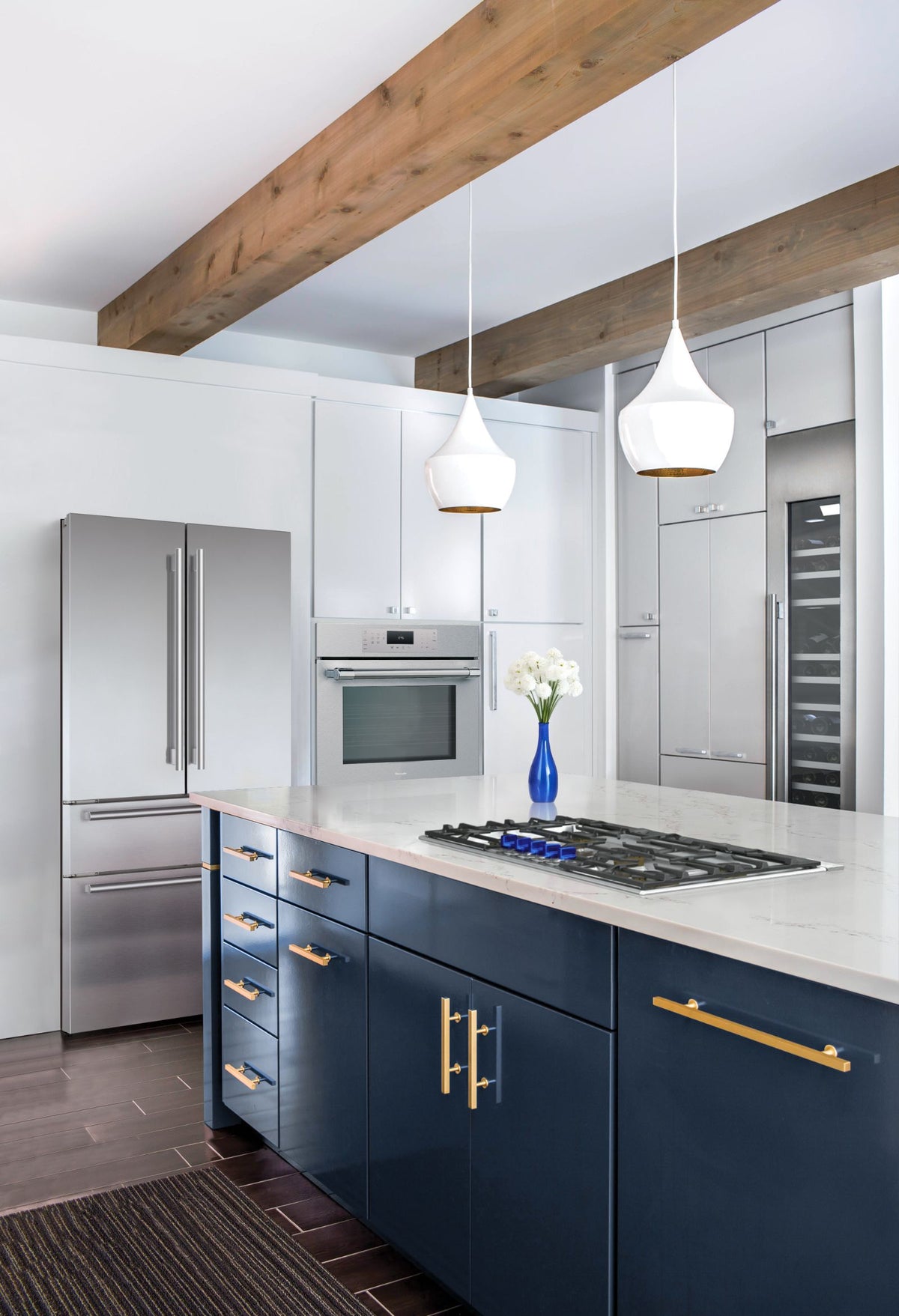 A modern kitchen interior featuring navy blue lower cabinets with gold handles, a white countertop, Thermador stainless steel appliances, pendant lights, and exposed wooden ceiling beams.