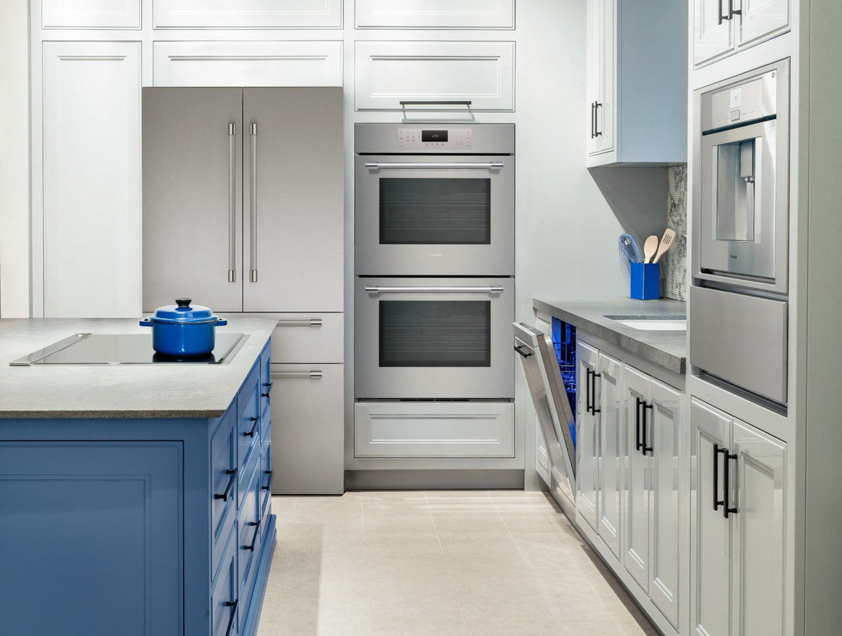 A modern kitchen interior with white shaker-style cabinets, Thermador stainless steel appliances including a side-by-side refrigerator and double wall oven, dishwasher, in-wall coffee machine, a blue island with a blue pot, and light beige tile flooring.