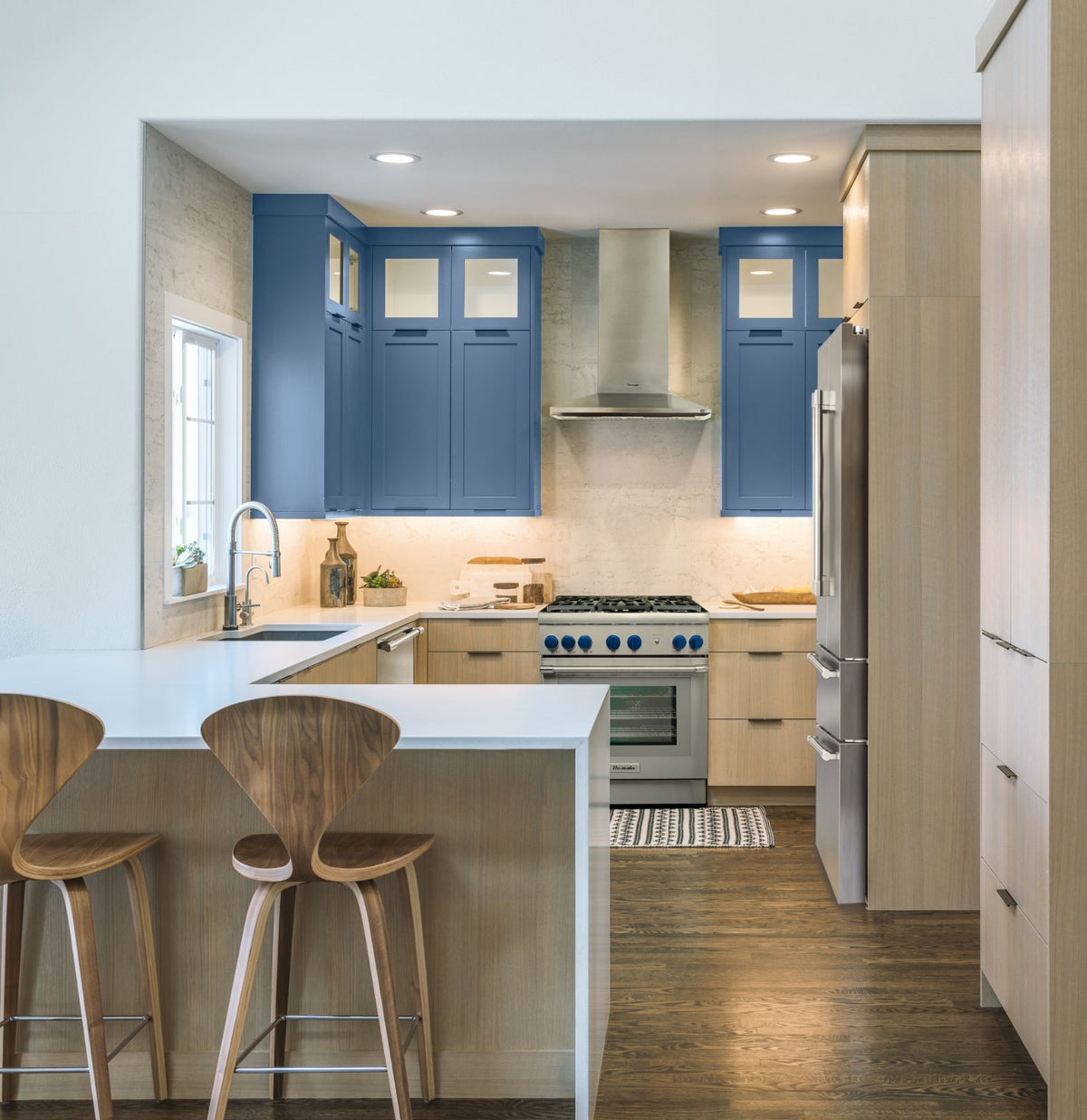 A modern kitchen interior with blue upper cabinets, light wood lower cabinets and island, white countertops, Thermador stainless steel appliances, and two wooden bar stools at the island.
