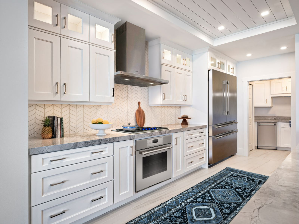 A modern kitchen interior with white shaker-style cabinets, Thermador stainless steel appliances including a refrigerator, dishwasher, oven, and range hood, a patterned tile backsplash, marble countertops, and a blue patterned rug on a light stone floor.